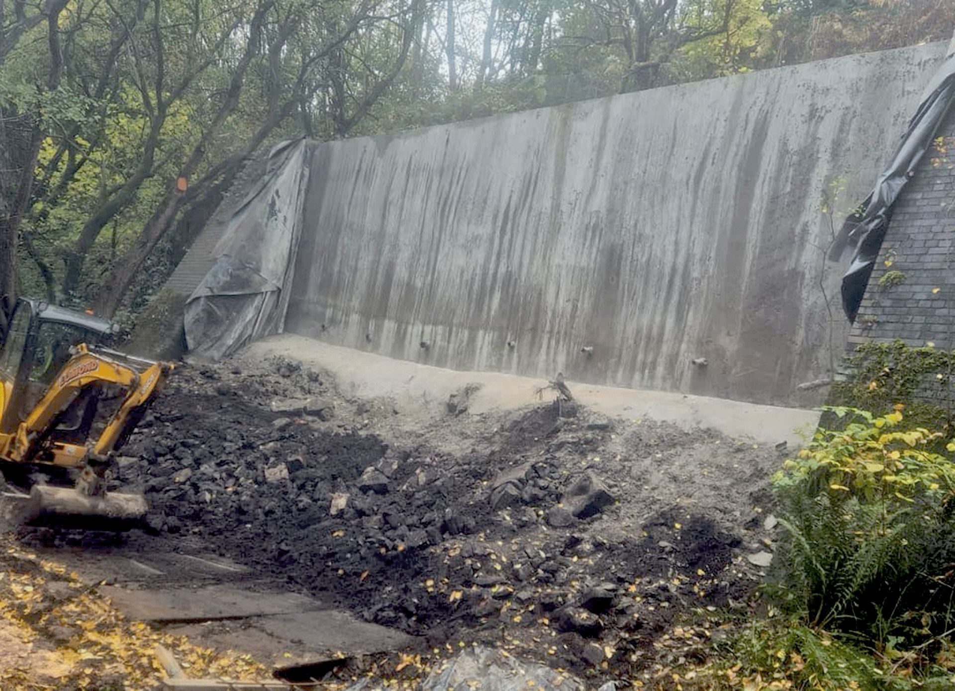 A roadside retaining wall with buttresses and rubble at the base and an excavator alongside.