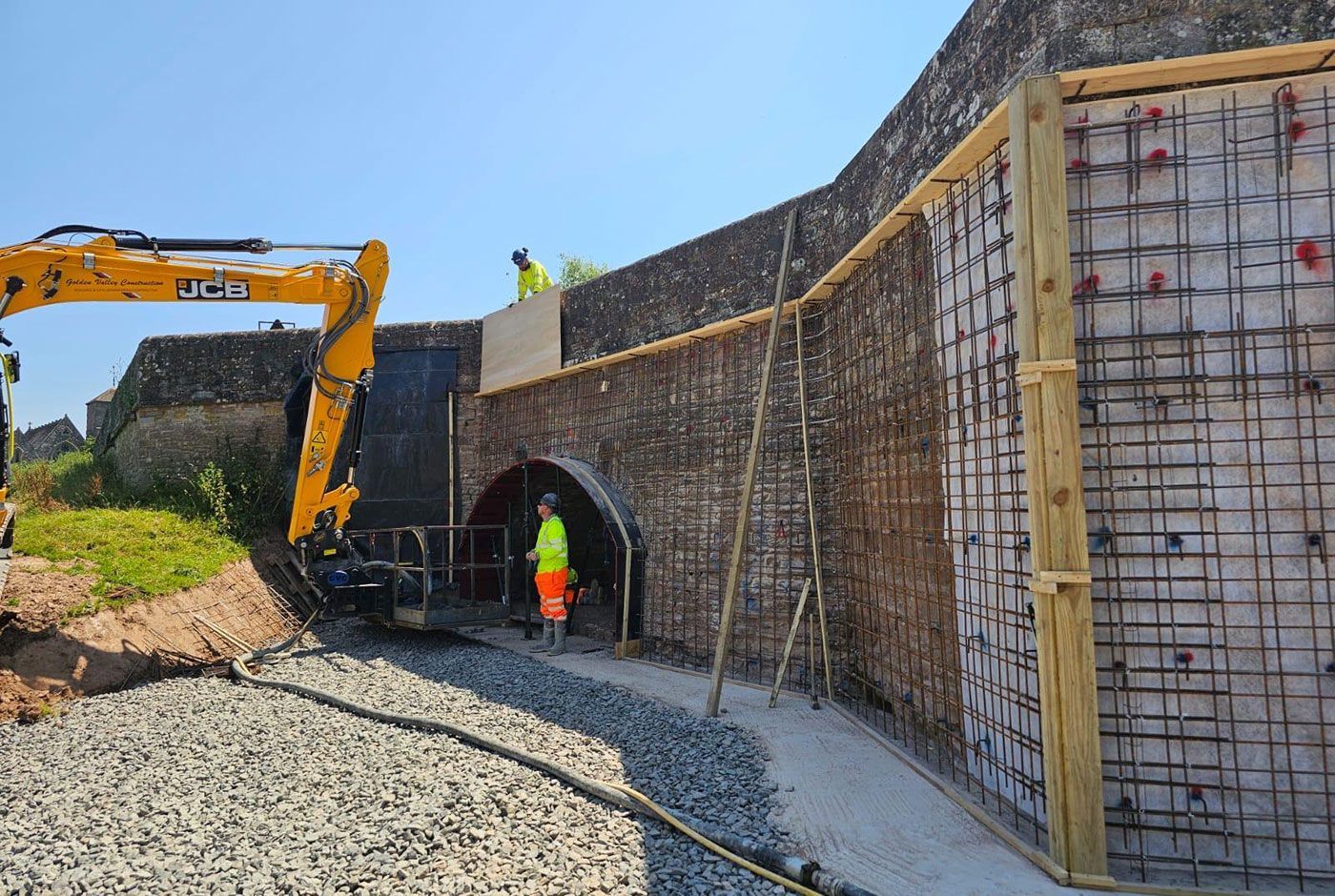 Construction workers at a stone bridge, repairs to the retaining wall being carried out, a JCB cherry picker alongside.