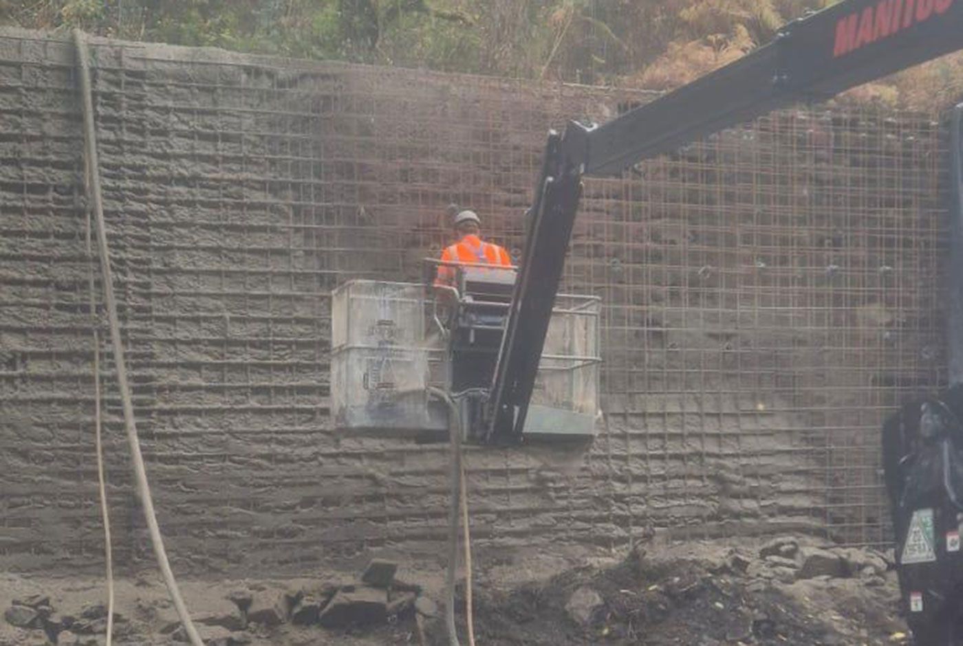 Construction worker in an orange vest in a cherry picker positioned against a wall under repair, mesh framework visible.