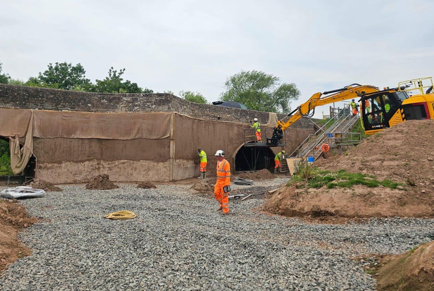Construction workers in hi-viz  at a bridge applying concrete to the retaining wall, a yellow cherry picker on the bank.
