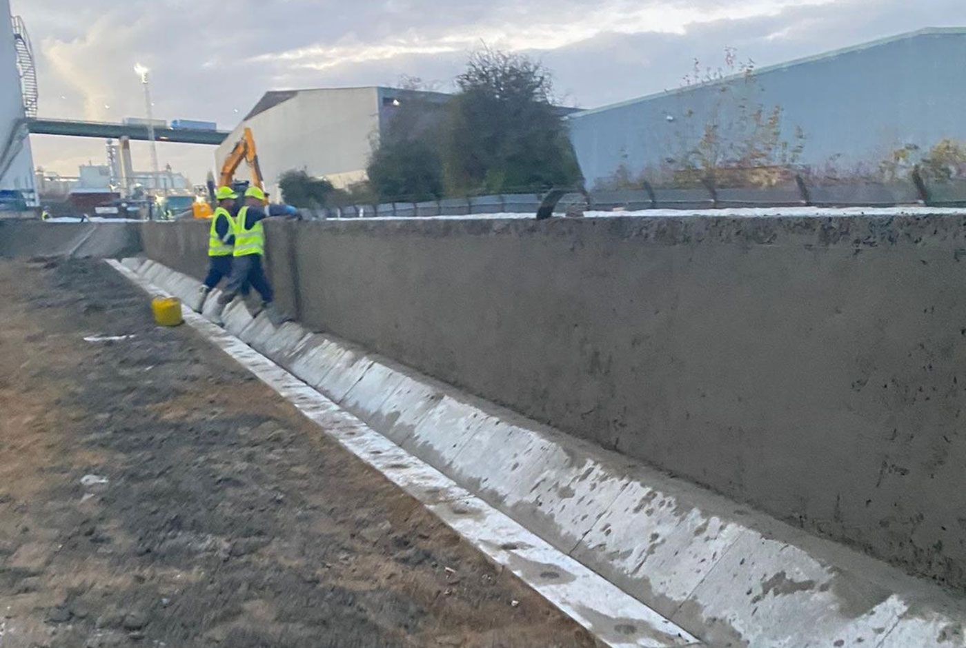 Workers in safety vests spreading concrete along a retaining wall on a roadside work site.