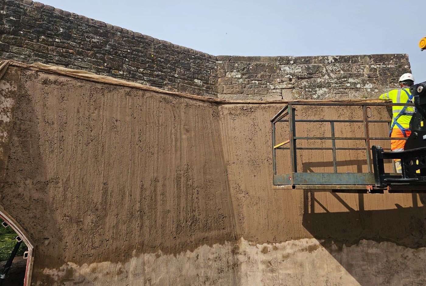 Worker in hi-viz gear on a platform working on a freshly concreted wall of a large stone wall.