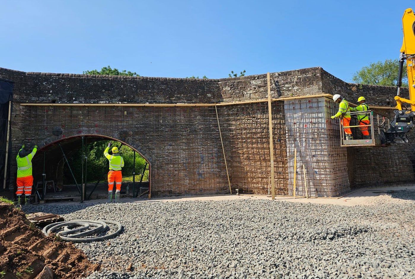 Workers in hi-viz gear repairing a stone bridge, preparing the walls with metal gridwork, blue sky above.