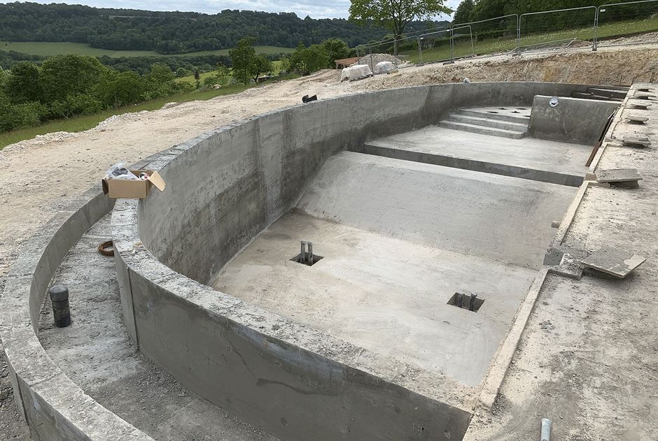 Concrete semi-circular pool shell under construction on a hillside, with stepped interior and surrounding gravel.