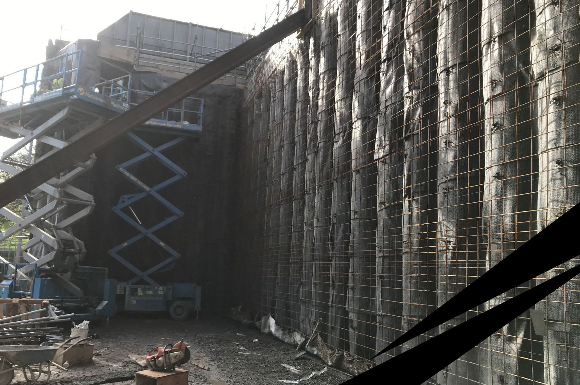 Construction site with tall concrete retaining wall and two scissor lifts fully extended beside it.