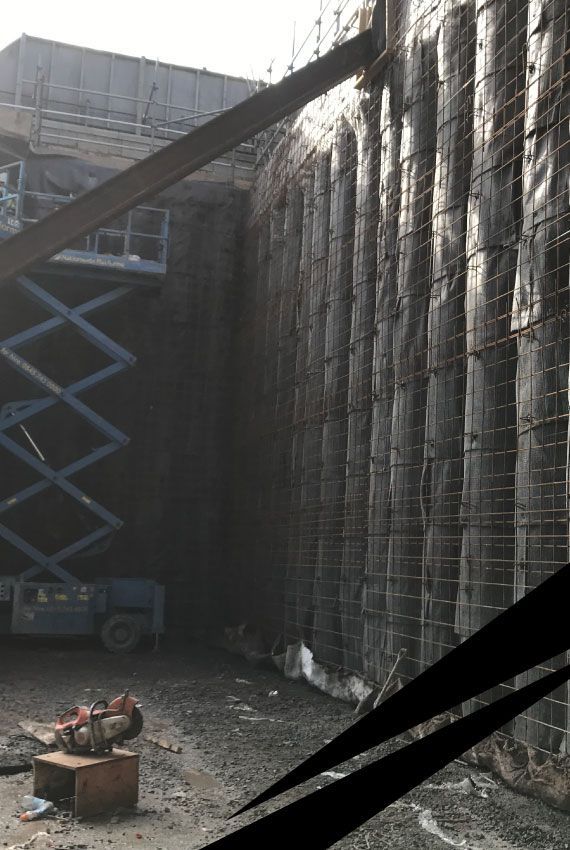 Construction site with tall concrete retaining wall and two scissor lifts fully extended beside it.