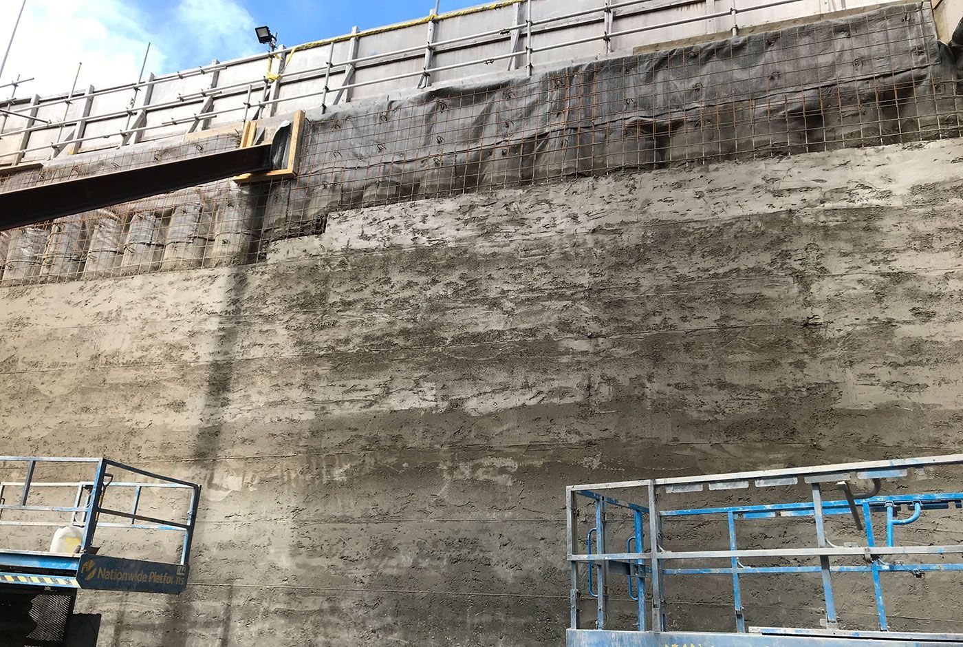 Concrete retaining wall under construction with blue scaffolding and safety railing at the top.