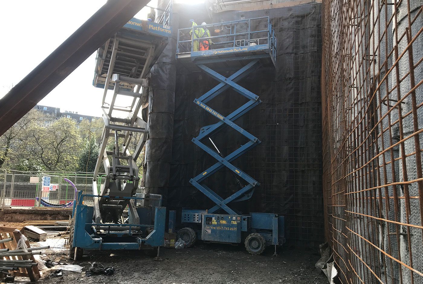 Two scissor lifts with their platforms raised beside a tall mesh-covered wall on a construction site.