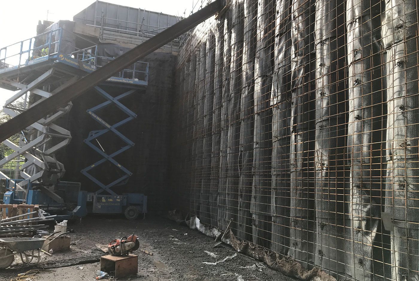 Construction site with tall concrete retaining wall and two scissor lifts fully extended beside it.