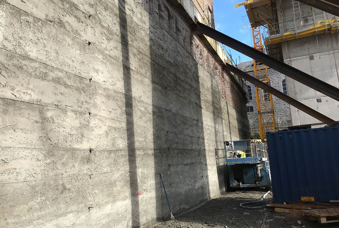 Construction site with tall concrete wall, steel beams, and a blue scissor lift.