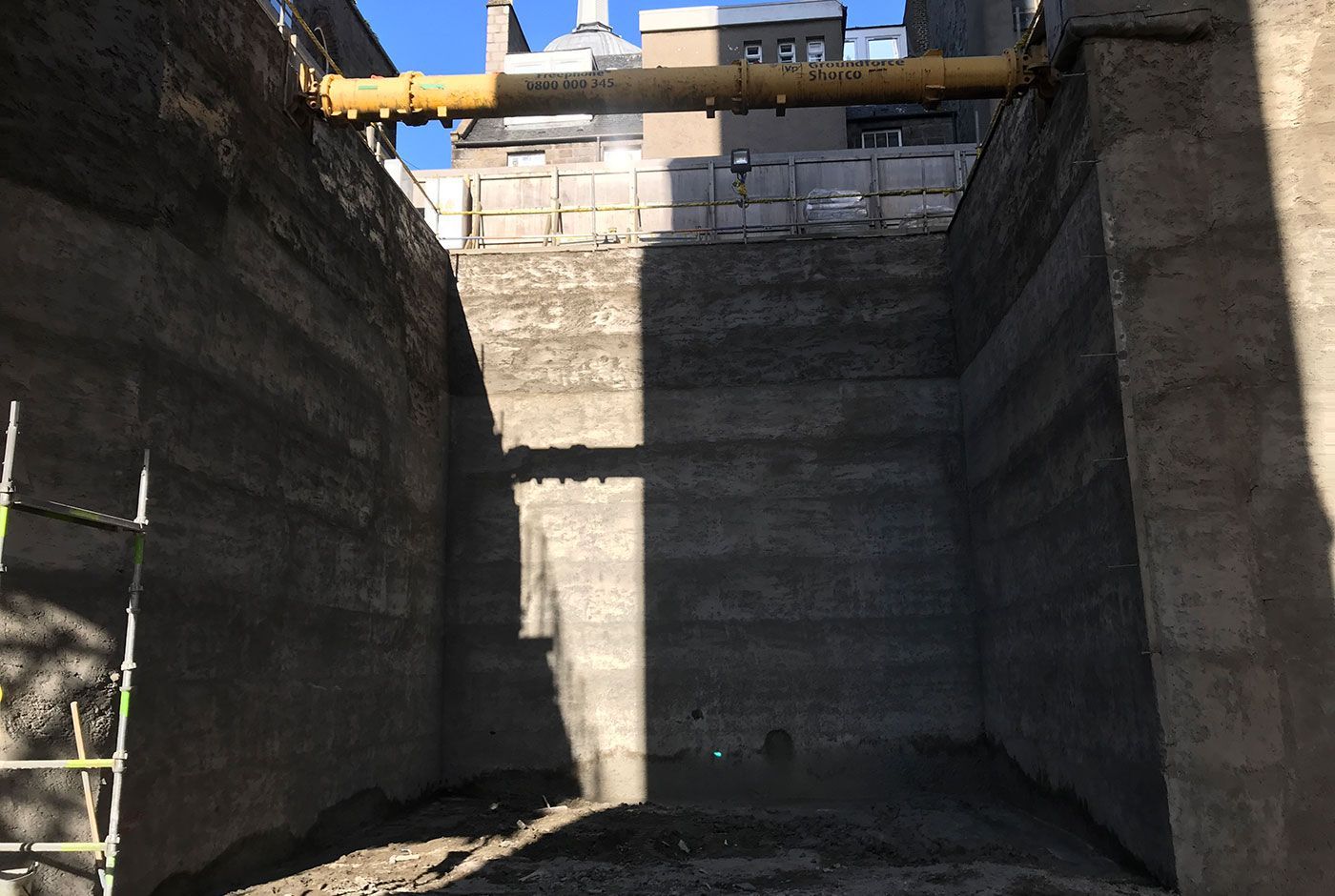 Construction site with tall concrete retaining walls and a yellow pipe overhead, lit by sunlight.