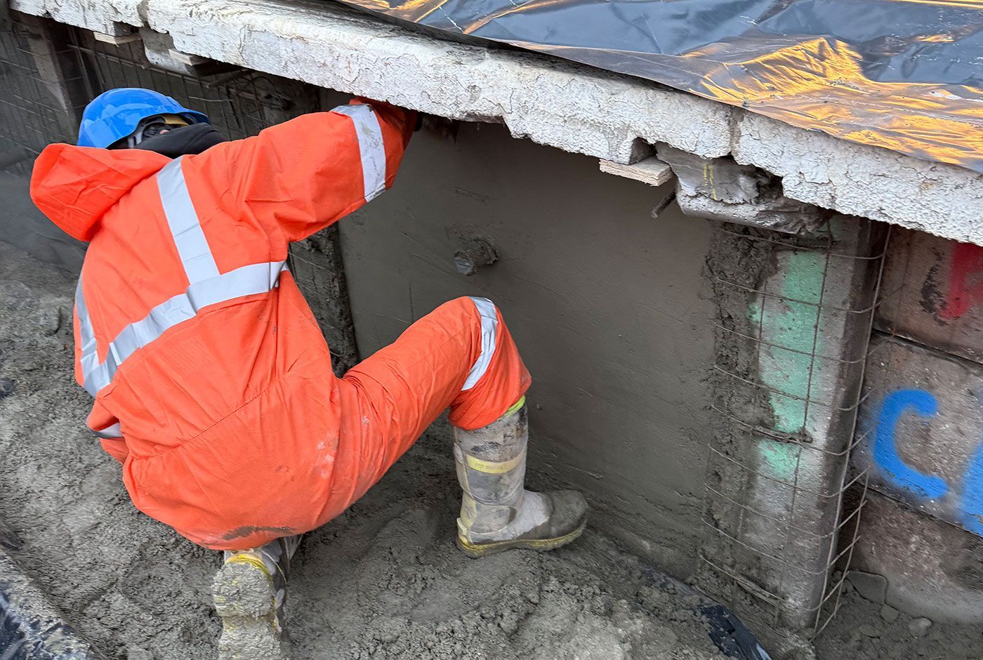 Worker in orange PPE crouches under a railway platform, applying fresh concrete to the platform wall.