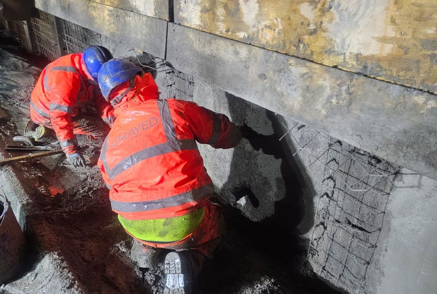 Two workers in orange PPE and blue safety hats crouch by a concrete wall, applying concrete repairs.
