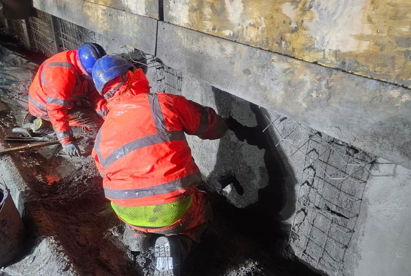 Two workers in orange PPE and blue safety hats crouch by a concrete wall, applying concrete repairs.