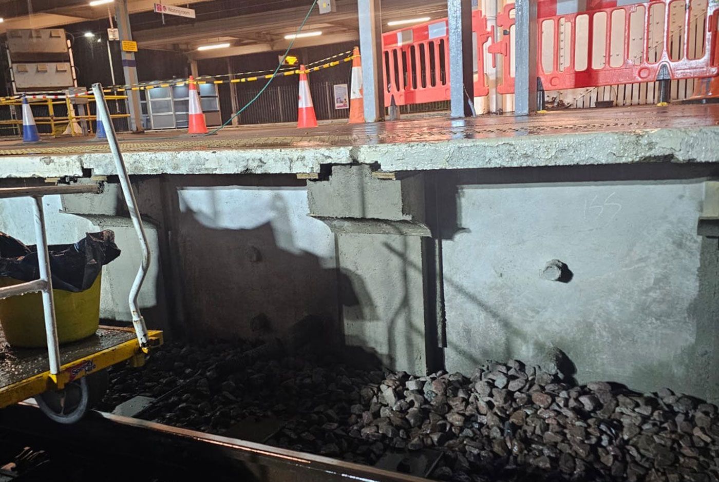 Damaged concrete wall of a railway platform under repair, with orange barriers and safety cones on the platform.