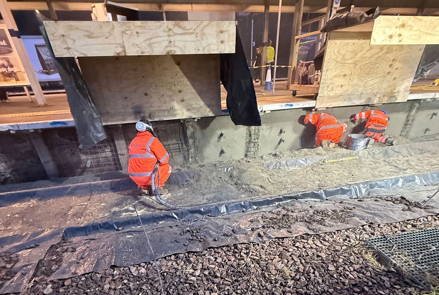 Workers in orange PPE working on concrete repairs to the wall of a railway platform, one is spraying concrete.