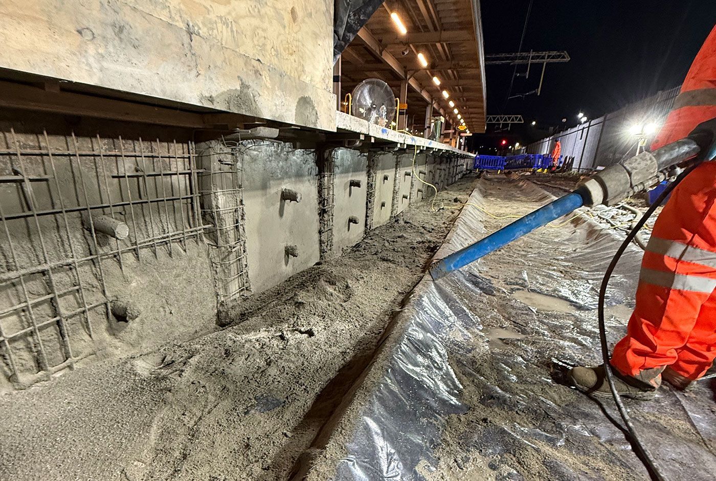 Railway platform wall repairs at night, with mesh on the wall and a worker in orange PPE holding a spray concrete nozzle.