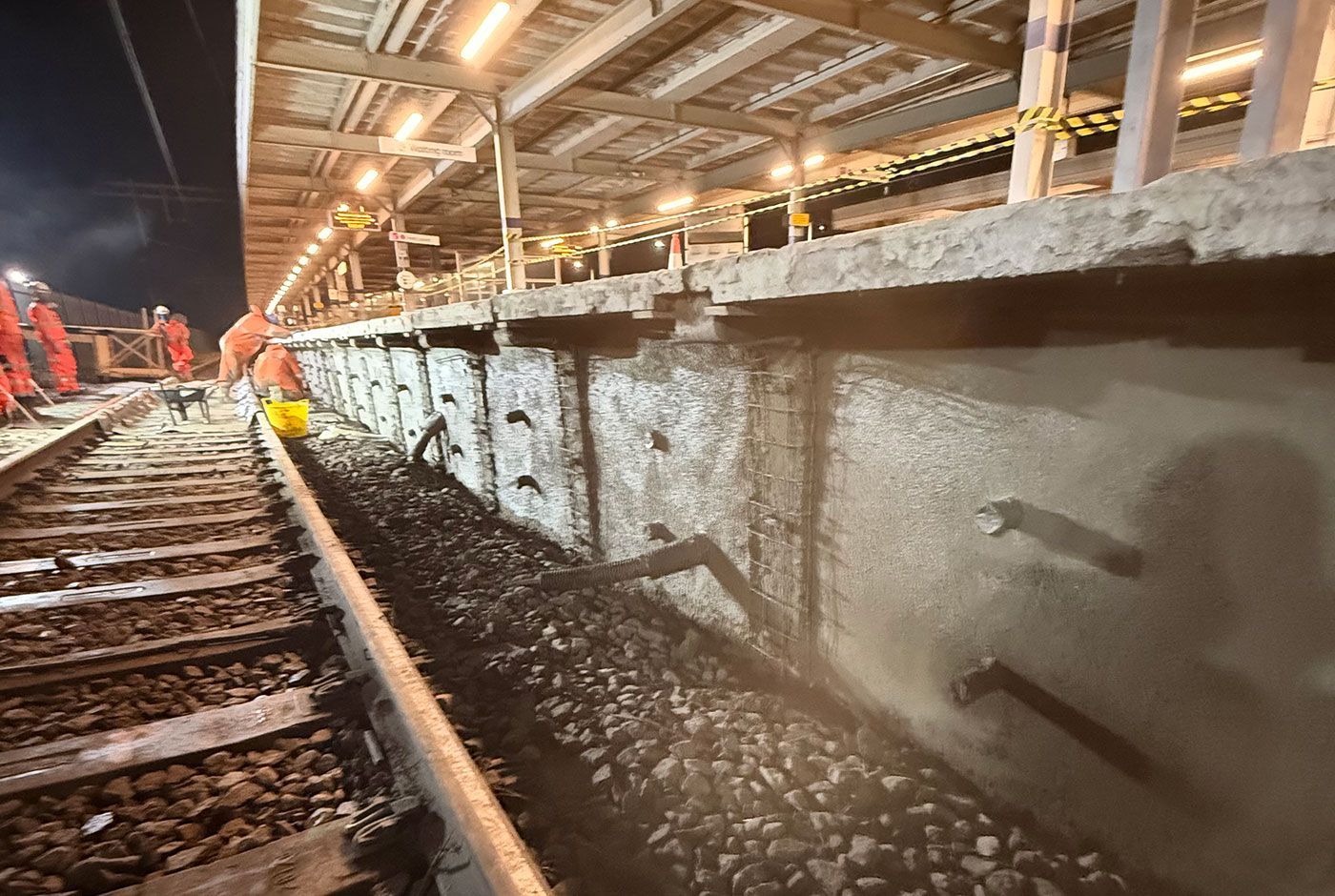 Construction workers on a lit nighttime railway platform beside a concrete wall and railway tracks.