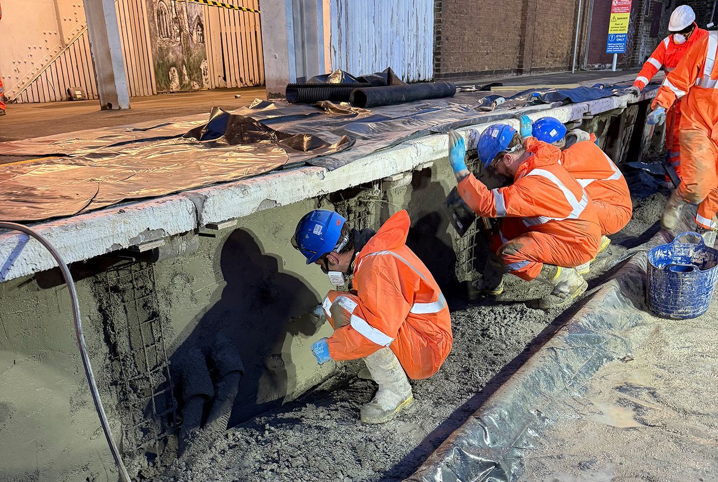Workers in orange safety gear and blue helmets making repairs to a railway platform wall.