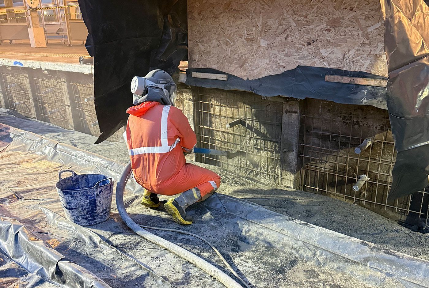 Worker in orange safety gear kneeling on a concrete surface repairing a railway platform wall  using sprayed concrete.