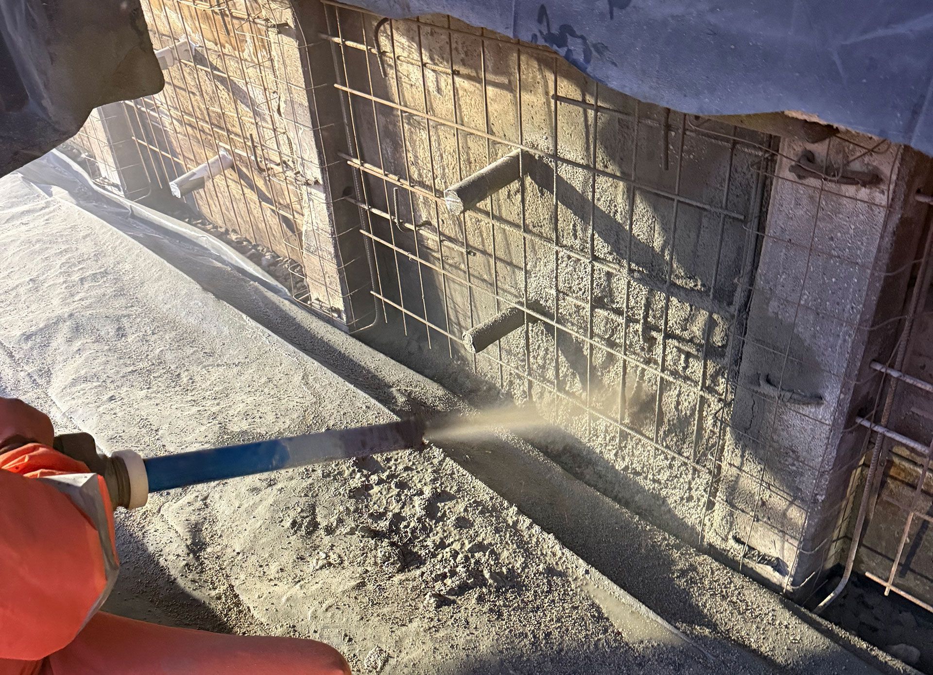 Construction worker holding a hose and applying sprayed concrete to a mesh-reinforced wall.