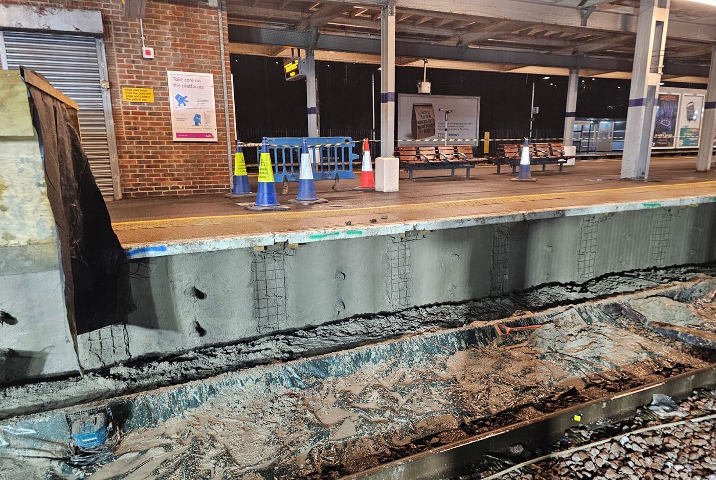 Railway platform, its retaining wall under repair with fresh concrete, rail tracks covered with tarpaulin and debris.