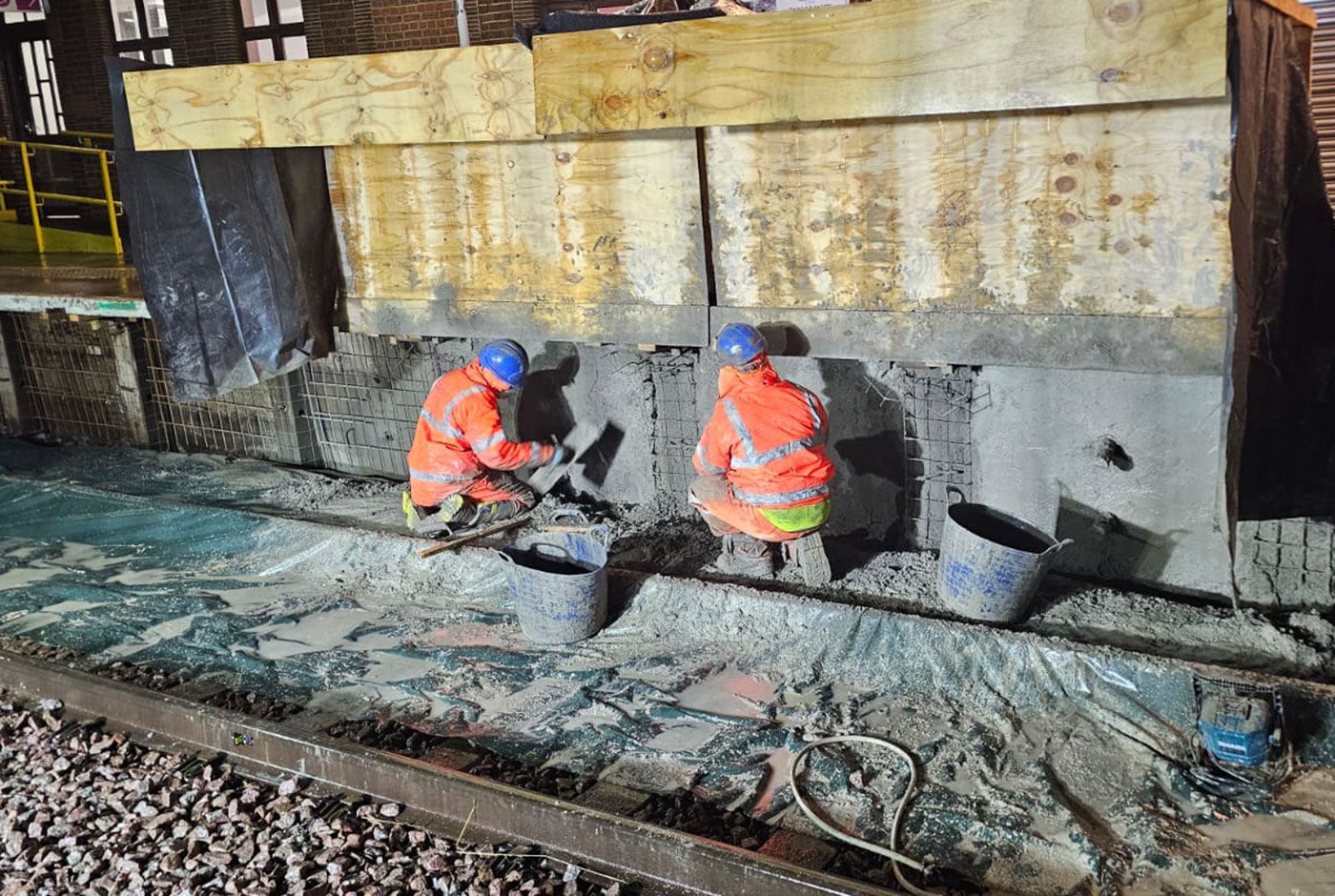 Two workers in orange PPE and blue safety helmets applying wet concrete with a trowel to a retaining wall.