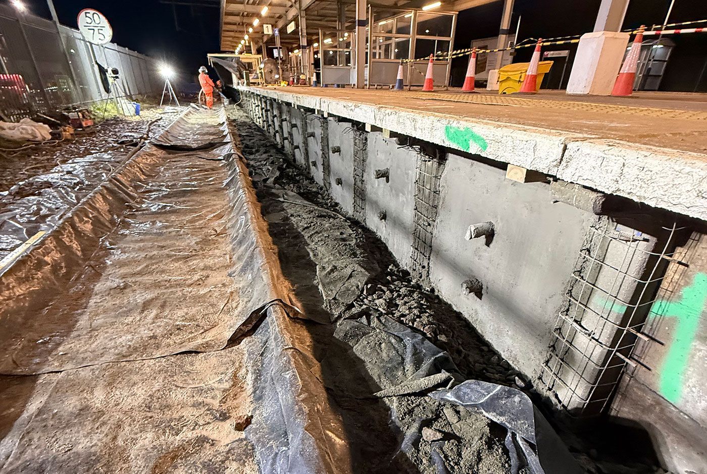 Night construction at a railway platform, with floodlight, concrete retaining wall, rail tracks covered with tarpaulin.