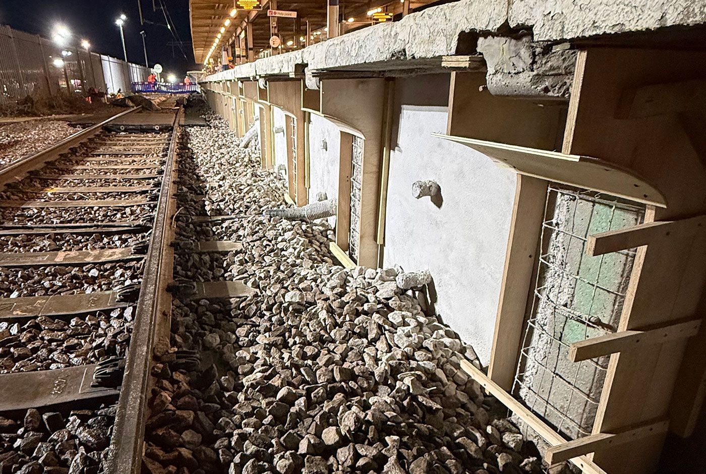 The wall of a railway platform at night, under construction with wooden formwork, rail track and heavy gravel.
