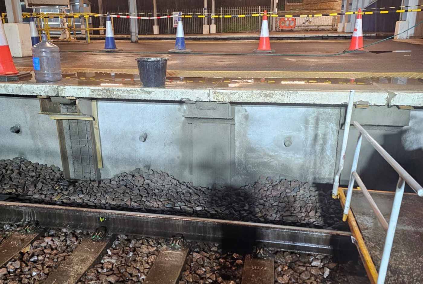Railway platform under repair with freshly applied concrete on the retaining wall, safety cones on the platform.