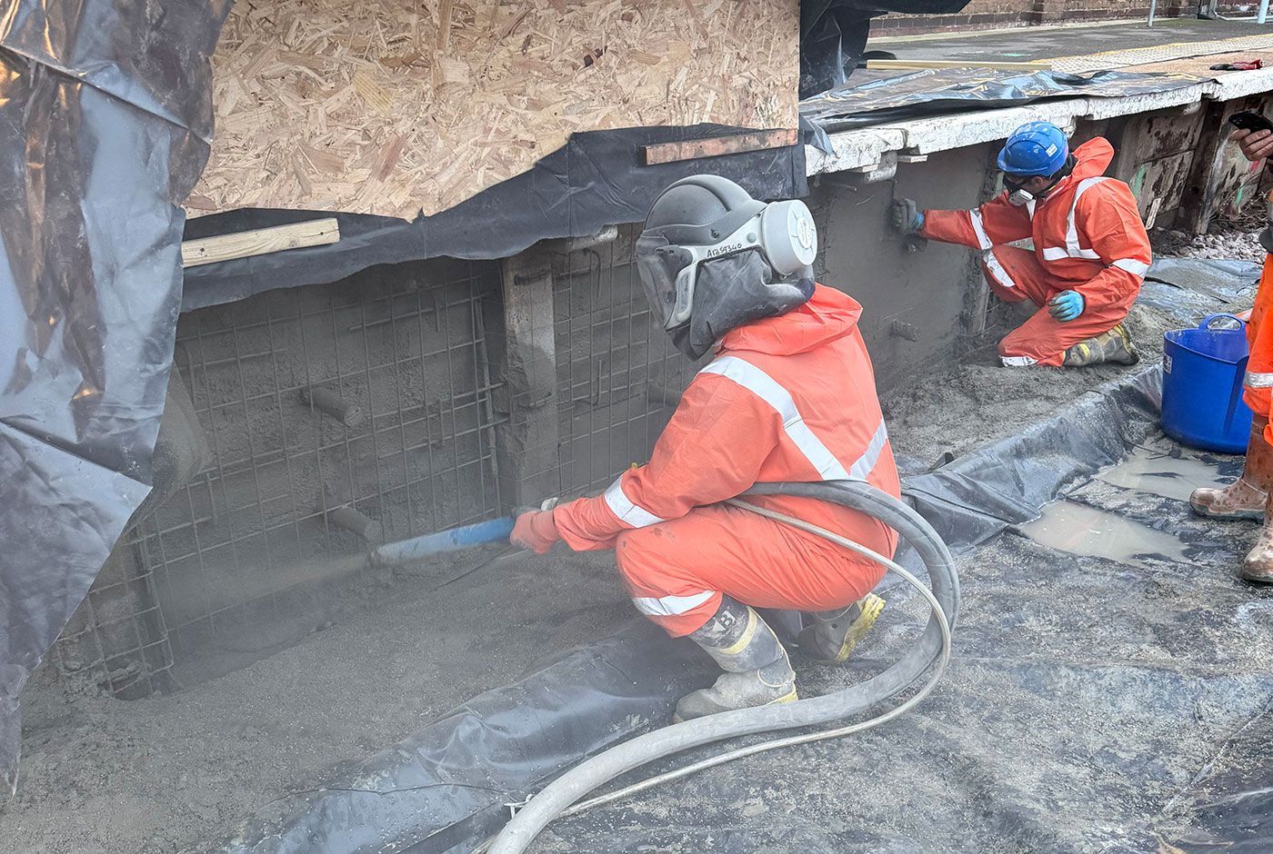 Two workers in orange PPE at a railway platform wall, one spraying concrete onto a mesh frame, the other smoothing concrete.