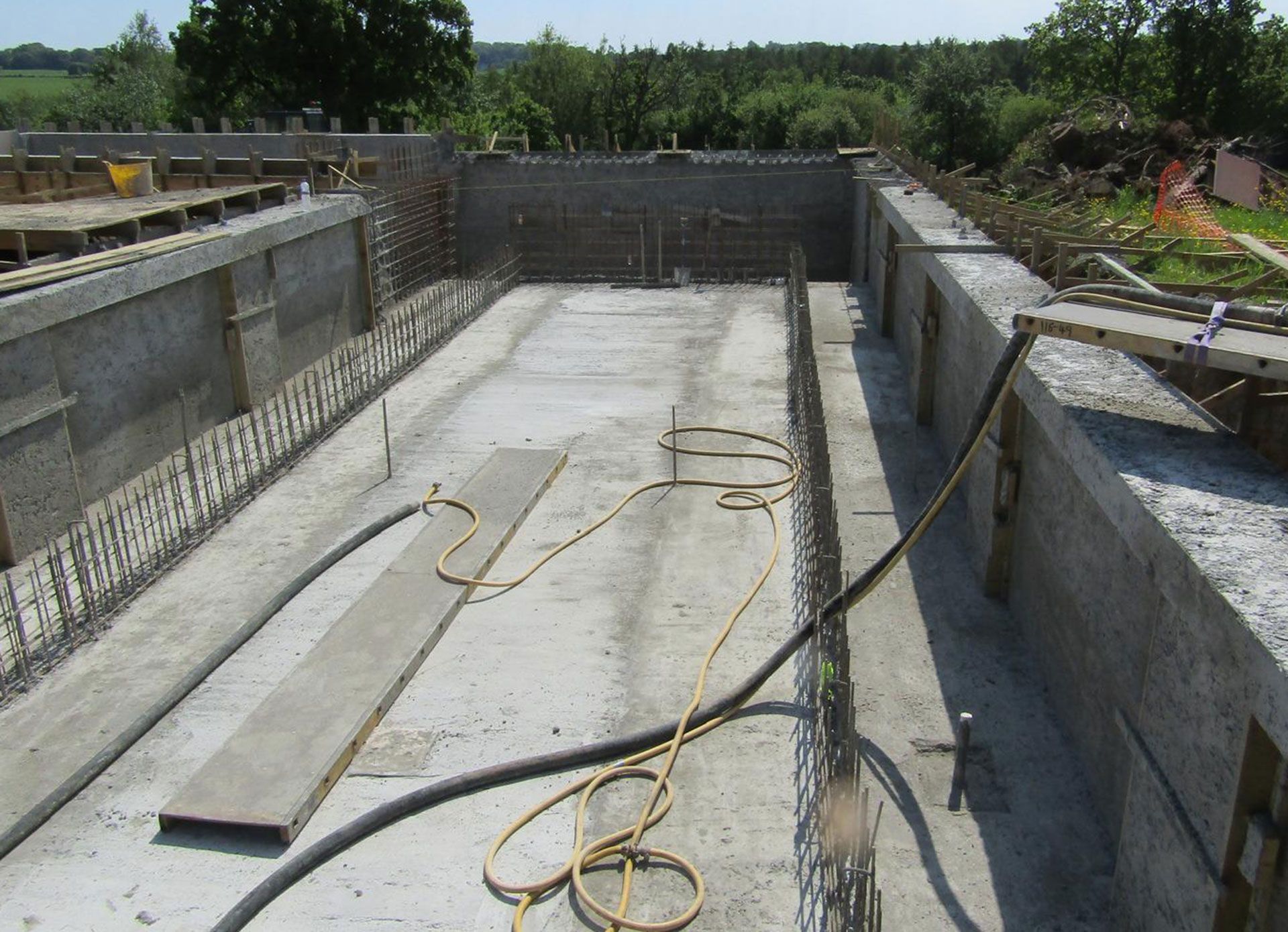 Open air concrete foundation under construction with various hoses on the floor, trees in the background.