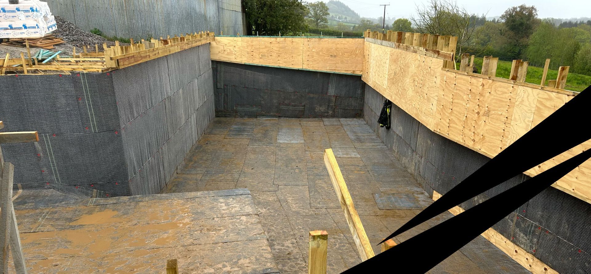 Concrete foundation site with wooden formwork and concrete floor under construction, countryside in the background.