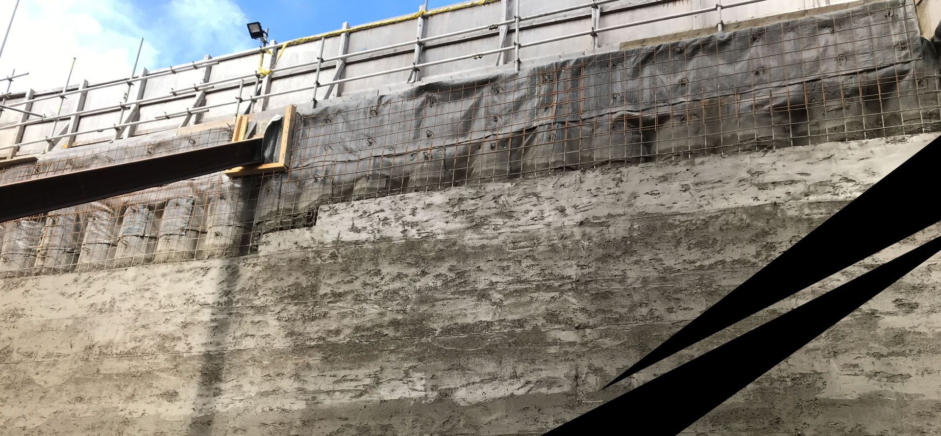 Contiguous wall with partly completed pile facing, barriers along the top and a bright sky above.