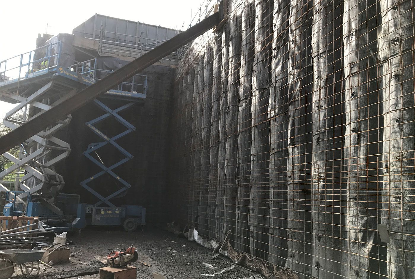 Two fully extended scissor lifts beside a tall wall covered with metal gridwork.