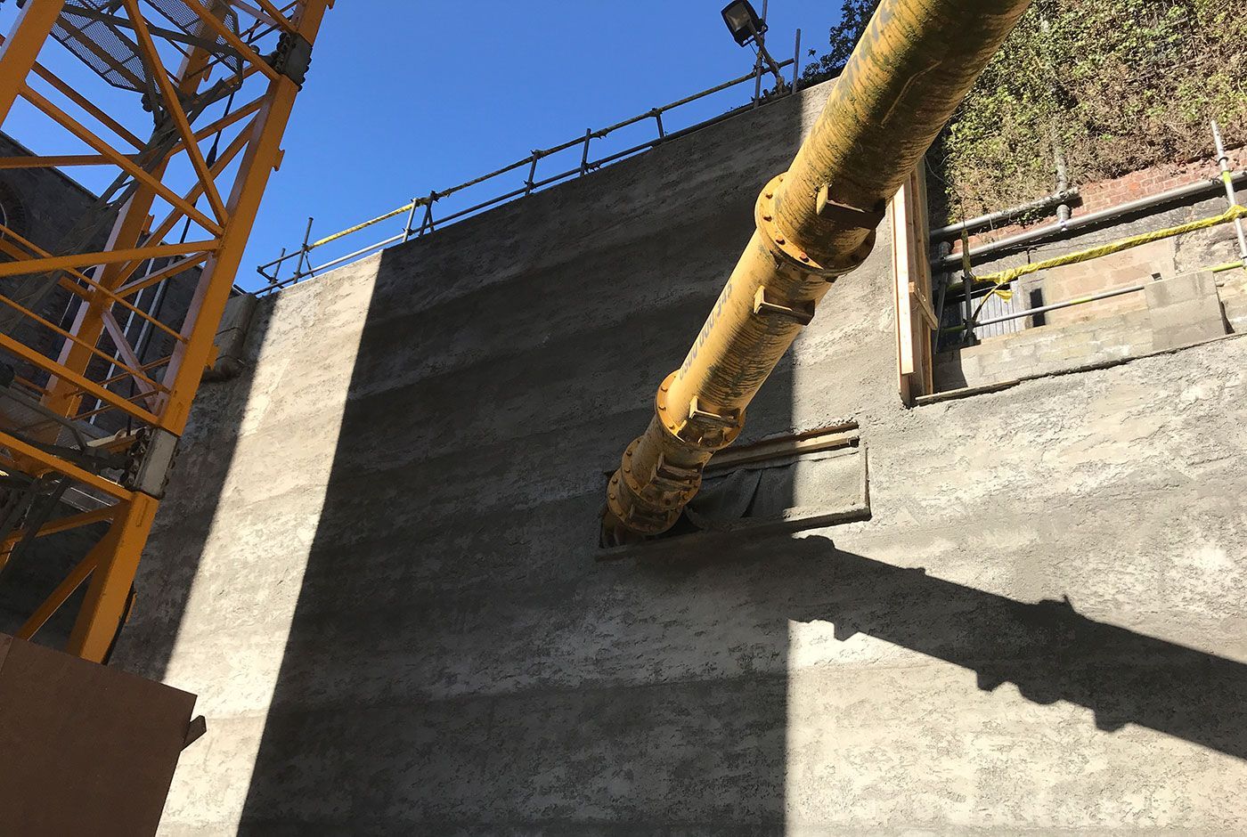 Concrete wall with a yellow pipework, part of a crane visible and blue sky above.
