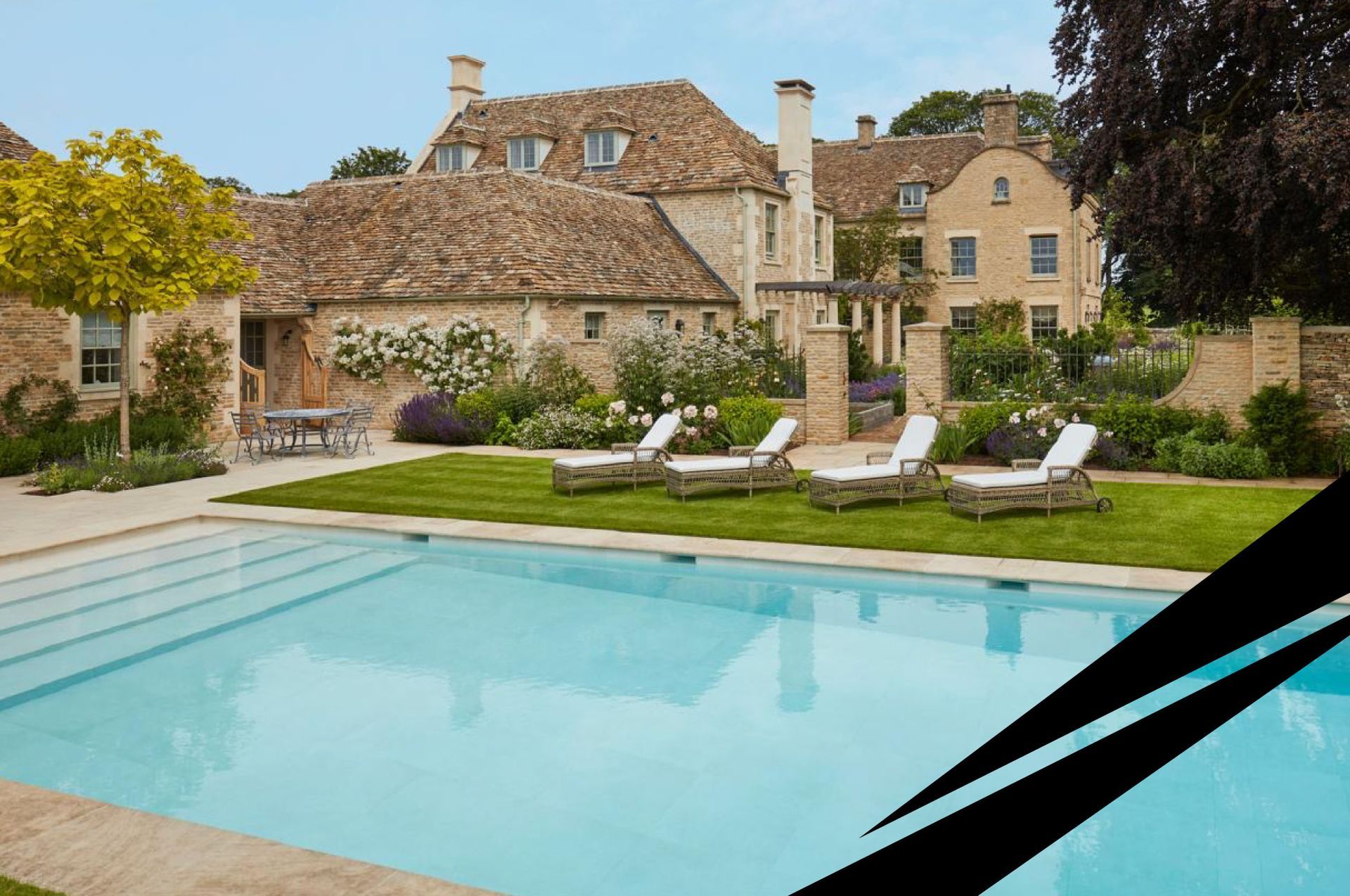 Garden view of a large stone house with a turquoise pool, lawned area and sunbeds in the foreground.