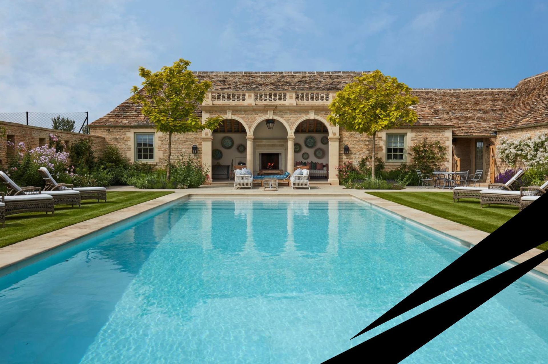 A country house courtyard with stone arches, a luxury turquoise swimming pool in the foreground with sunbeds on either side.