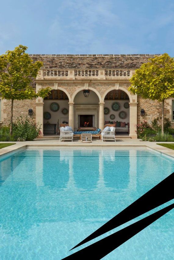 A country house courtyard with stone arches, a luxury turquoise swimming pool in the foreground with sunbeds on either side
