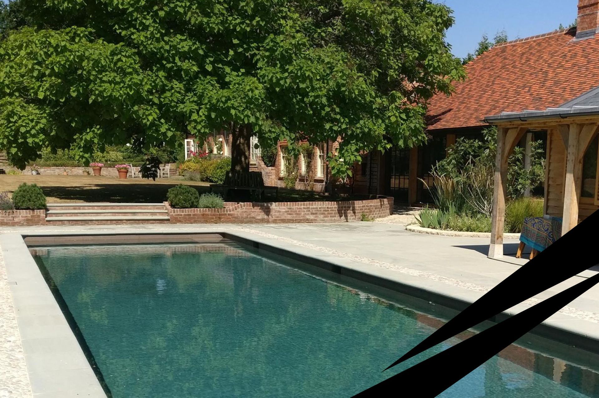 Residential swimming pool set within a patio with leafy trees and a red-tiled house in the background.