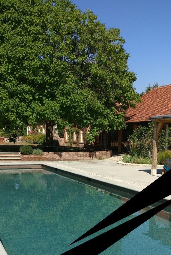 Residential swimming pool set within a patio with leafy trees and a red-tiled house in the background.