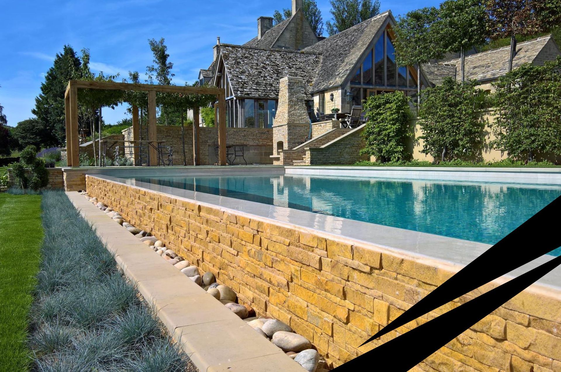 Stone house with steep roof with a turquoise swimming pool and landscaped greenery in the foreground.