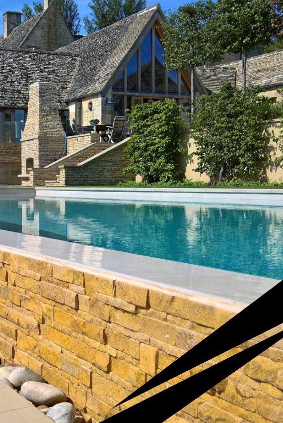 Stone house with steep roof with a turquoise swimming pool and landscaped greenery in the foreground.