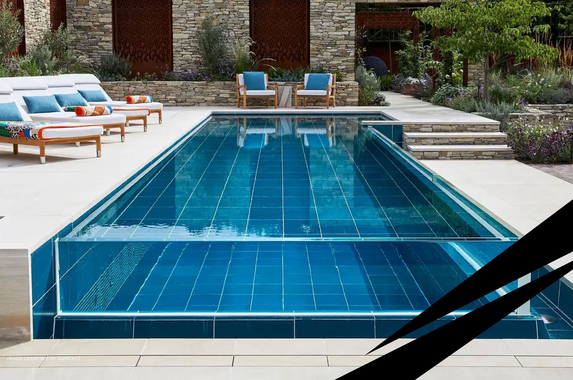 Modern outdoor pool with blue water and glass wall, patio area with steps, sunbeds, against a brick wall