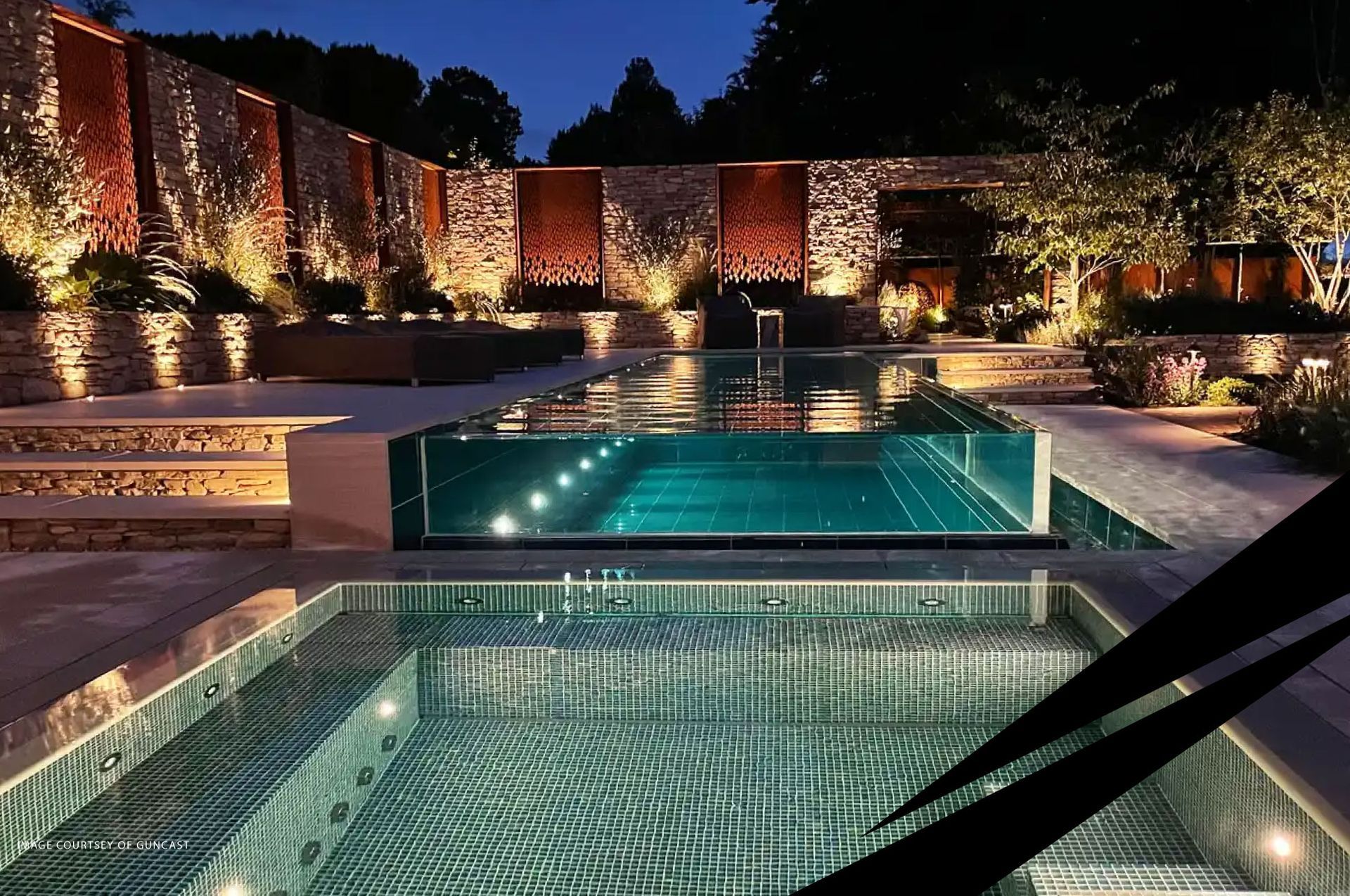 Illuminated modern pool terrace at night with reflecting water, stone walls, and surrounding greenery.