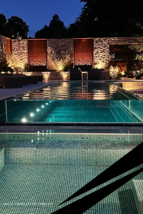 Illuminated modern pool terrace at night with reflecting water, stone walls, and surrounding greenery.