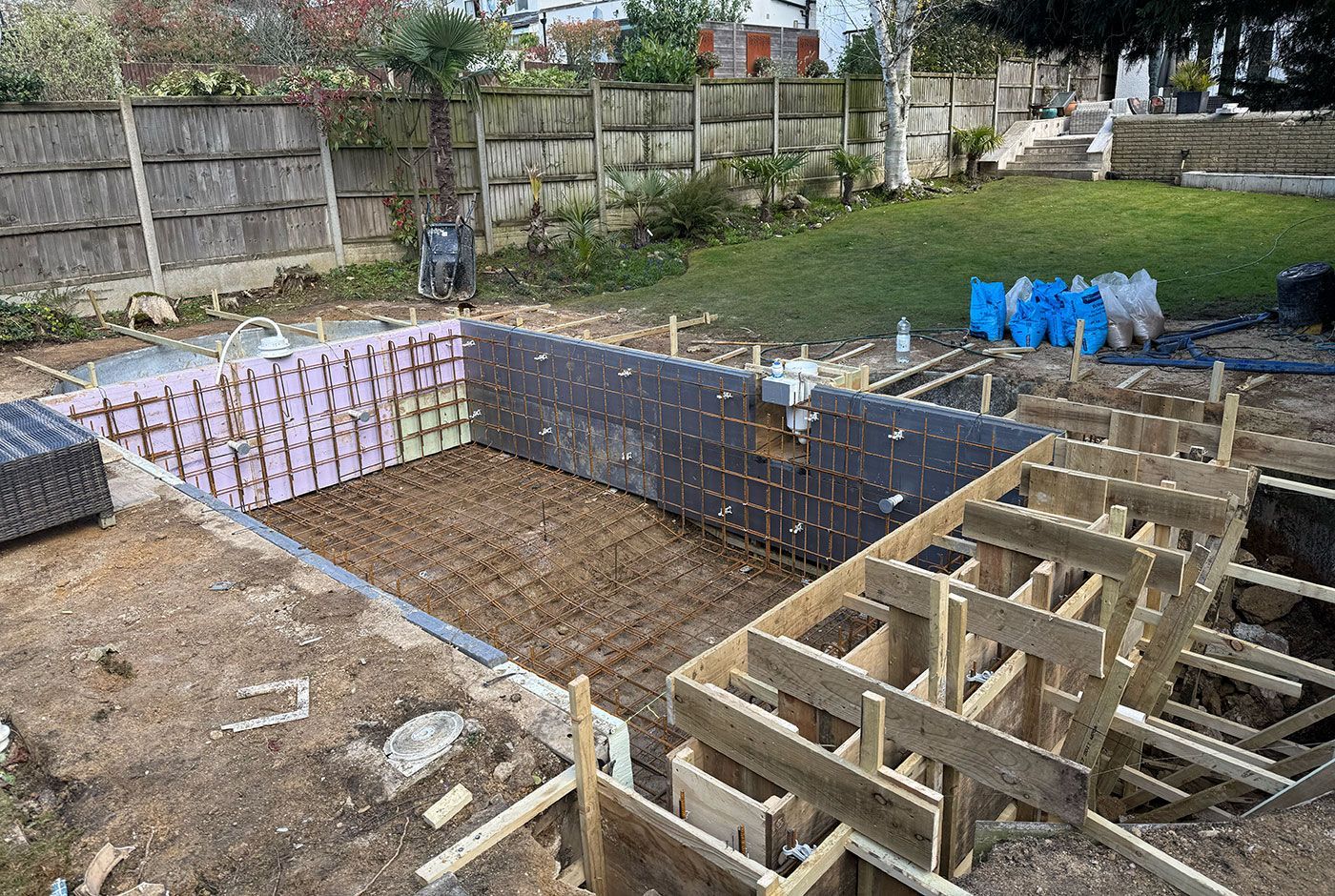 Construction of a swimming pool with wooden formwork, rebar, tools and bags by a lawned area.