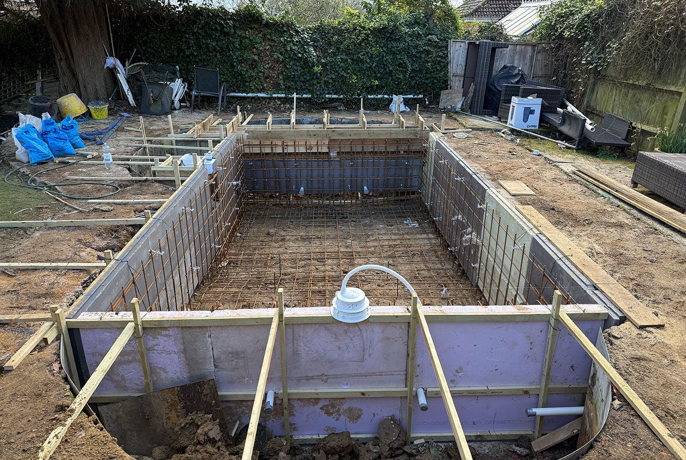 Framework of a swimming pool foundation in a garden, with wooden supports and construction materials around it
