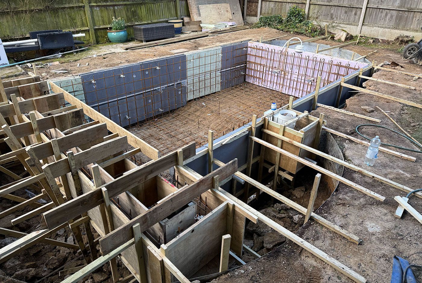 A rectangular swimming pool in construction, with wooden formwork in a garden, panel fencing in the background.