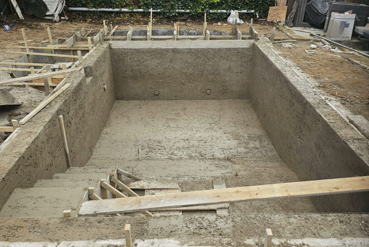 Concrete swimming pool shell under construction, wooden braces against steps, and unfinished surroundings.